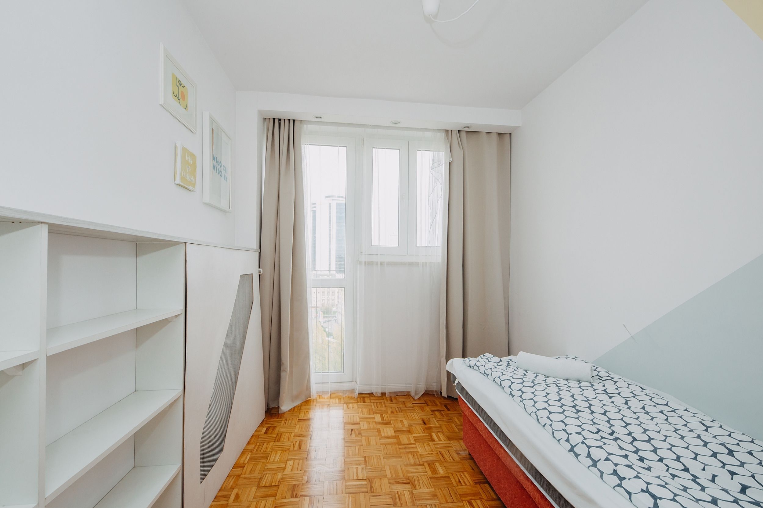 Una habitación pequeña y luminosa con piso de madera, una cama con ropa de cama estampada, un armario blanco y una ventana con cortinas.