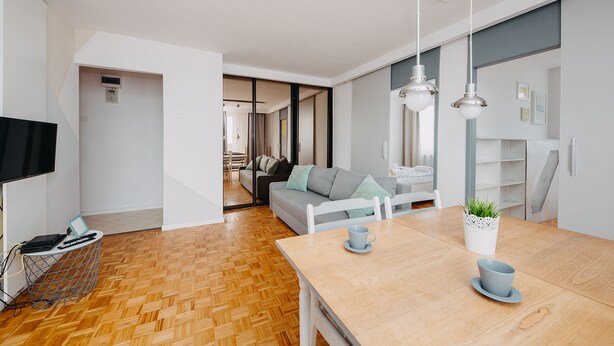 Una sala de estar moderna con piso de madera, un sofá gris, una mesa de comedor con dos sillas y un televisor a la izquierda.
