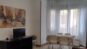 Een woonkamer met een flatscreen televisie op een zwarte standaard, een eettafel met stoelen, een bruine bank en een groot abstract schilderij aan de muur.