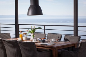 Une table à manger agrémentée de chaises en osier, un vase rempli de fleurs et une vue sur la mer par de grandes fenêtres.