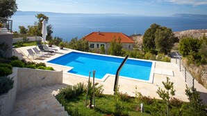 En swimmingpool med ett rött tegeltak, omgiven av en stenmur och grönska, med utsikt över ett vackert havsscenarium.
