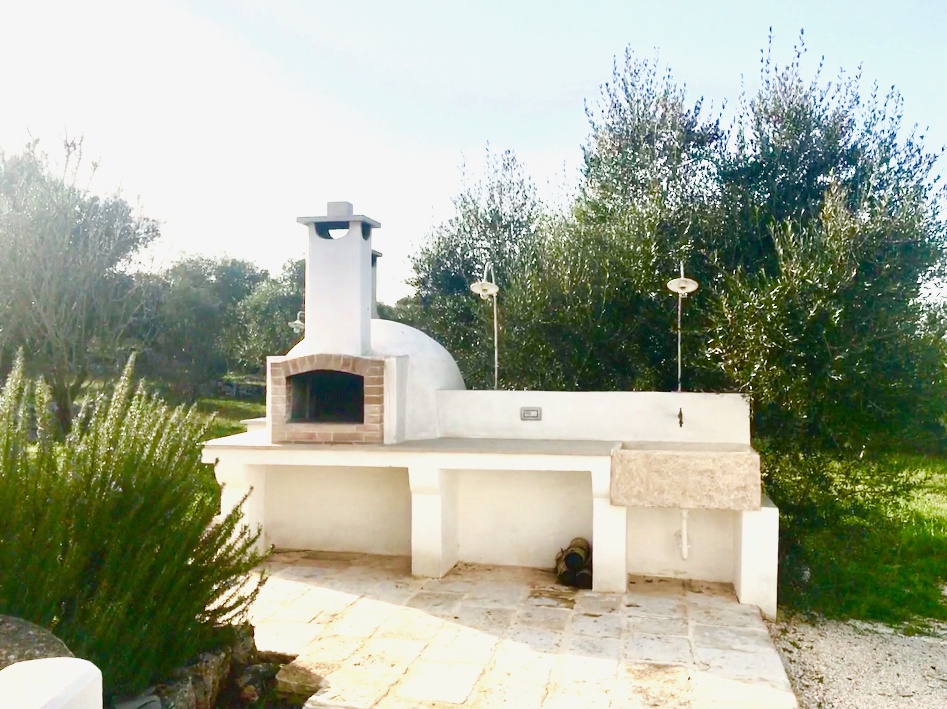 Un four à pizza extérieur blanc, doté d’une cheminée, entouré de verdure et bordé d’une zone pavée.