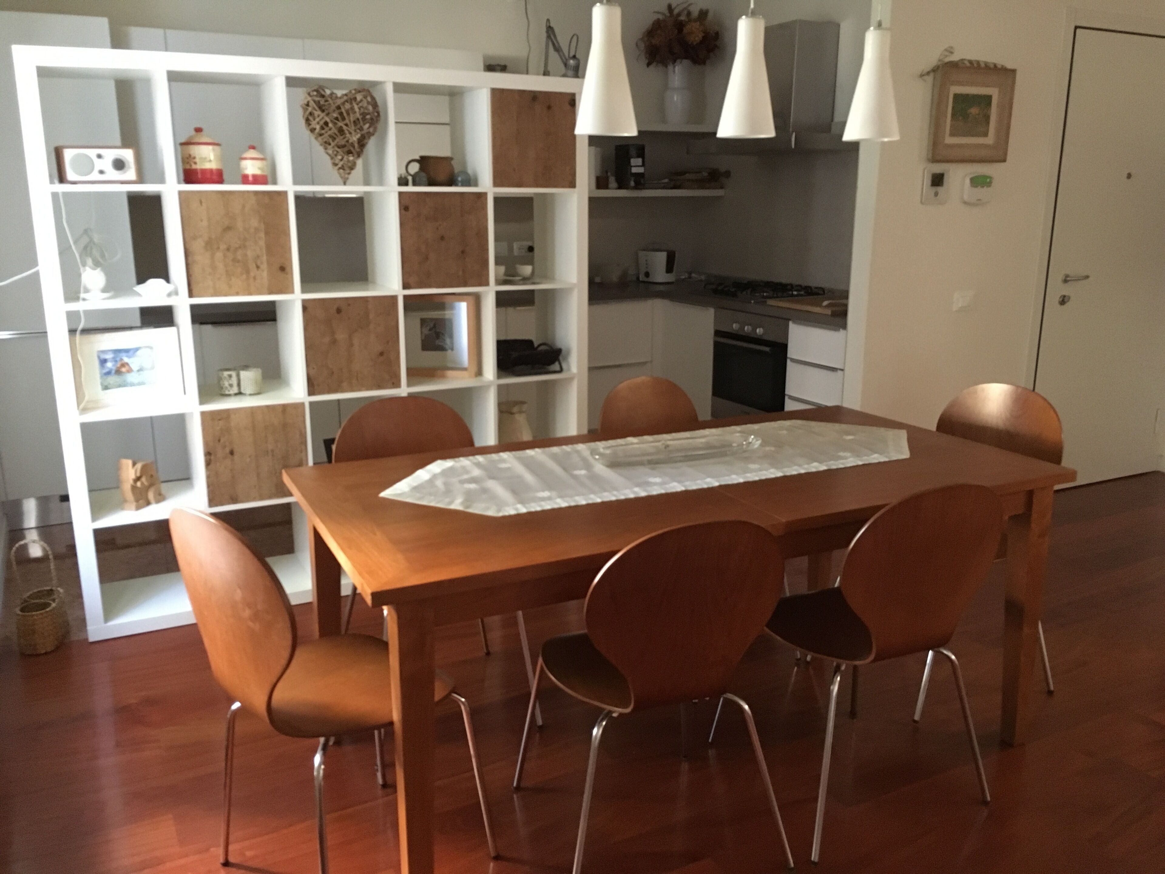 A dining area with a wooden table and six chairs, a white shelving unit with various items, and a kitchen in the background.