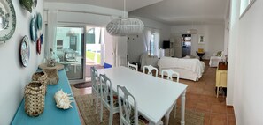 A spacious living area with a dining table, white chairs, a sofa, and a kitchen in the background.