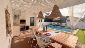 An outdoor patio with a wooden table, white wicker chairs, a pool, and a pergola with hanging lamps.
