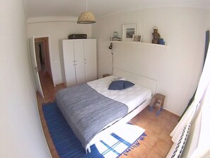A bedroom with a bed, a wardrobe, a hanging light fixture, and a small shelf with decorative items.