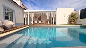 Una piscina con un porche cubierto, mobiliario de mimbre y una planta en maceta.