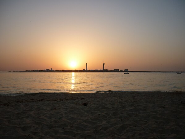 A beach with a sunset, calm water, and distant buildings.