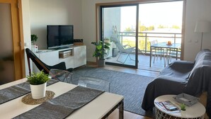 A modern living room with a flat-screen TV, a sofa, a dining table, and a balcony with outdoor seating. - Comfortable, cozy and comfy apartment by the Ria Formosa (Olhão)