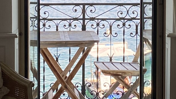 Un balcó amb taula i cadires, amb vista a una marina plena de vaixells i un paisatge amb muntanyes al fons.