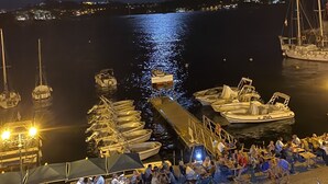 Una marina de nit amb vaixells amarrats, una vista clara de la lluna i gent dinovant a taules exteriors.