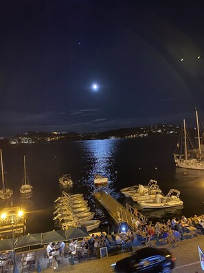 A marina at night with boats docked, a clear view of the moon, and people dining at outdoor tables.