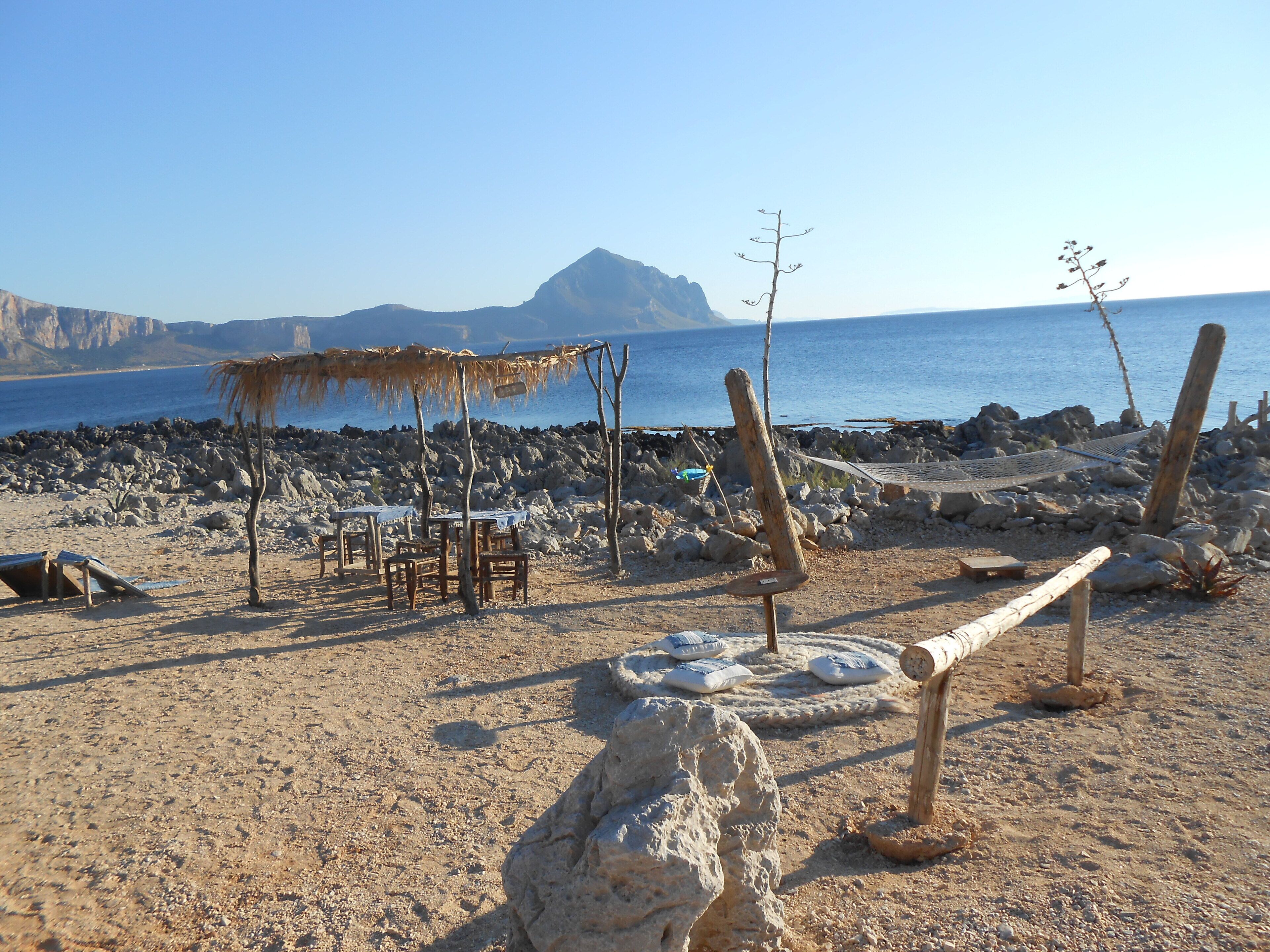 A beach with a table and chairs under a thatched umbrella, surrounded by rocks and a view of the sea and mountains.
