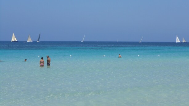 Une plage bordée d’une eau turquoise et cristalline, avec plusieurs voiliers visibles au loin.