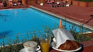 Une vue sur la piscine, avec des chaises longues et des parasols, une vue dégagée sur la mer, et une table avec une tasse de café, un verre de jus de fruits et un croissant.