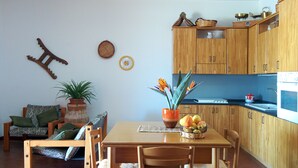 A compact kitchen with wooden cabinets, a dining table, and chairs. There is a potted plant on the table and a wall clock above the cabinets.