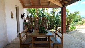 An outdoor patio with a wooden table and benches, surrounded by lush tropical plants and a wooden lattice fence.