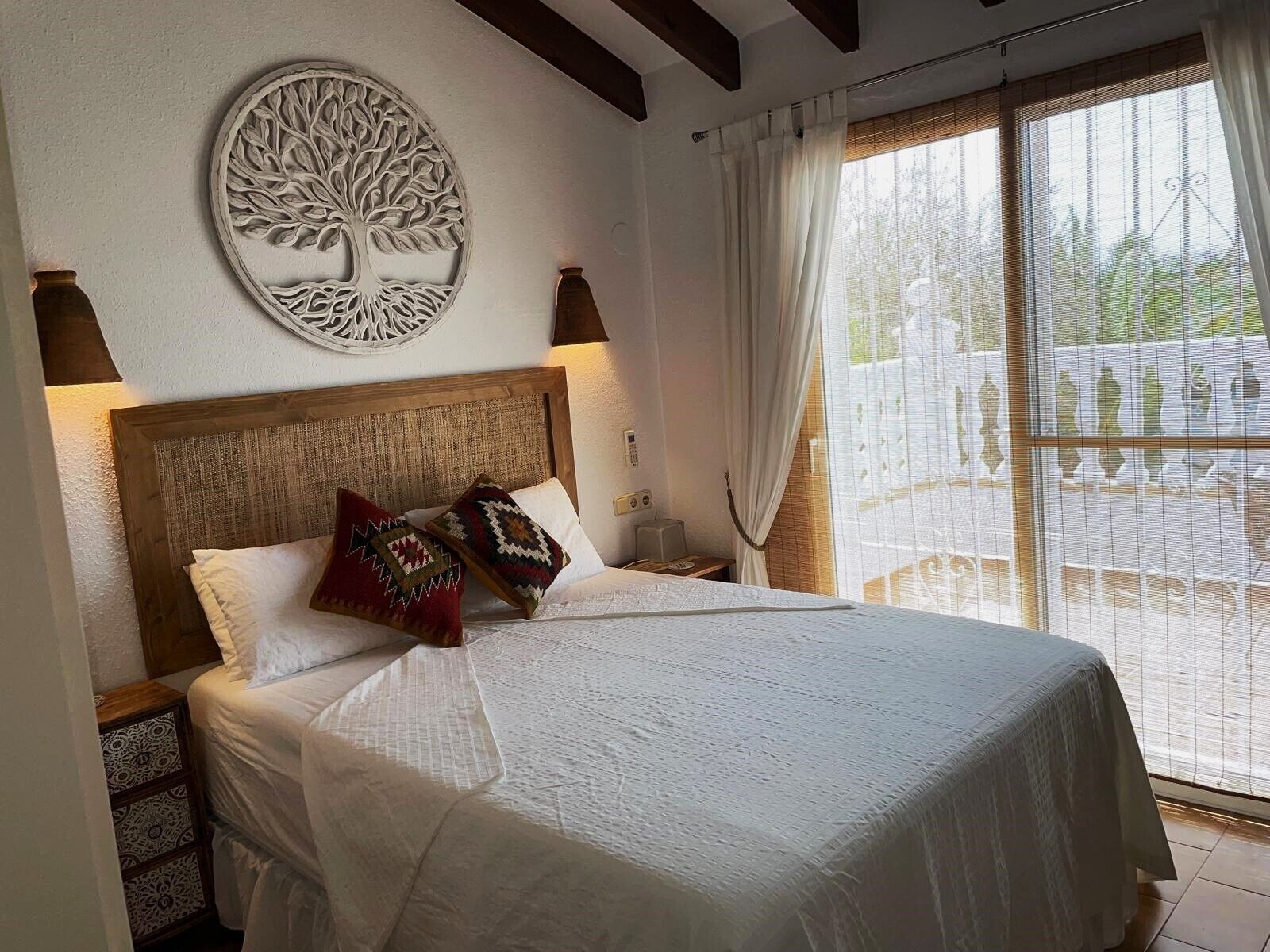 Une chambre à coucher avec une tête de lit en bois, un grand lit avec une literie blanche, deux coussins décoratifs, une sculpture d’arbre fixée au mur et une fenêtre avec des rideaux blancs.