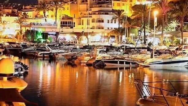 A marina at night with boats docked, a building with multiple balconies, and palm trees. - Villa Ra - Enchanting Moraira Villa with Secluded Pool and Sea View      (Alicante)