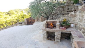 A stone-built outdoor barbecue area with a fire, a basket of vegetables, and a cutting board with meat.