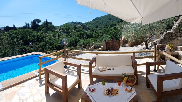 A poolside area with wooden chairs and a table set with snacks, overlooking a pool and a scenic hillside.