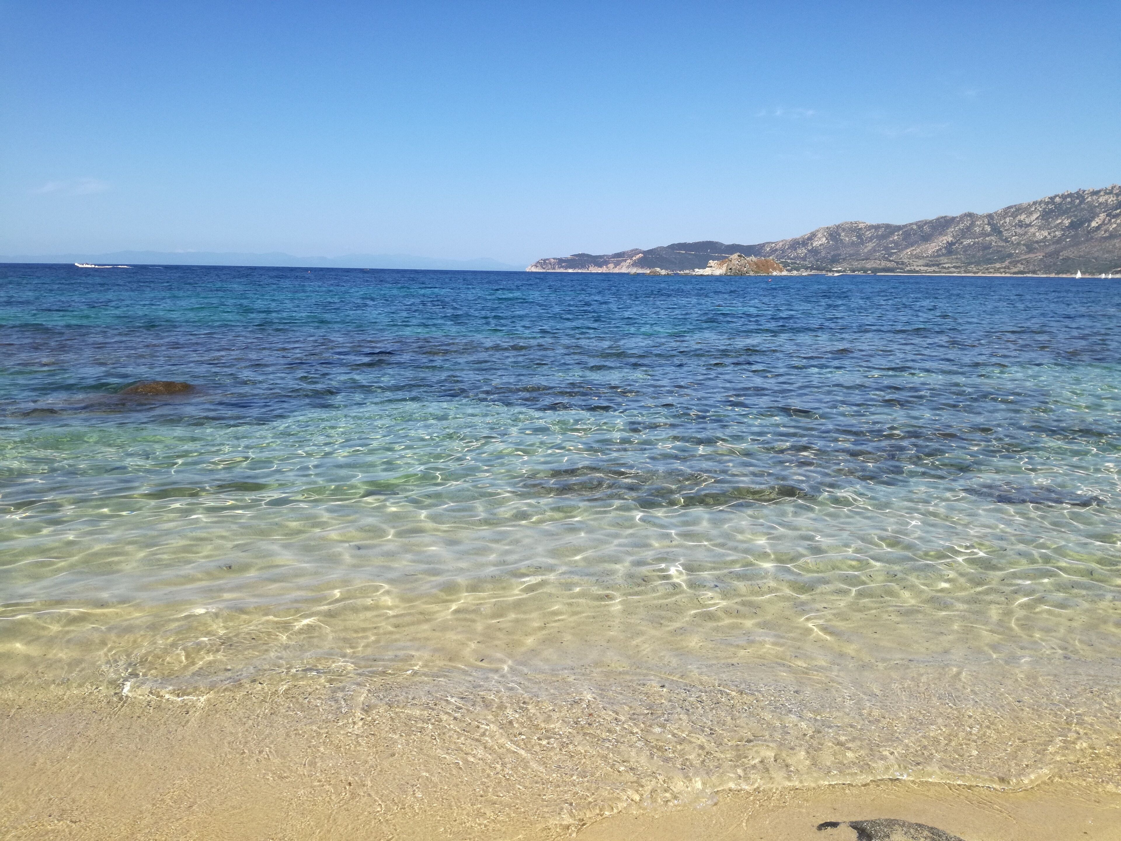 A beach with clear shallow waters and distant rocky hills.