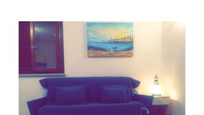 Une pièce avec un canapé bleu, un tableau représentant un paysage côtier, une fenêtre avec des stores fermés et une table de chevet avec une lampe.