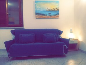 A room with a blue sofa, a painting of a coastal scene, a window with closed blinds, and a nightstand with a lamp.