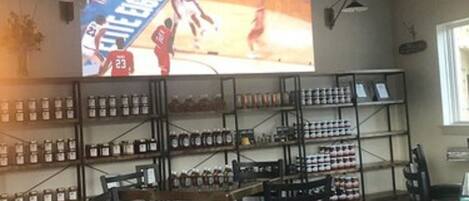Une pièce avec une grande télévision diffusant un match de basket-ball, des étagères remplies de bocaux et des chaises noires.