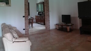 Un soggiorno con un divano beige, una TV su un mobile basso e una cucina visibile tramite un arco.