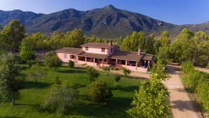 Una casa a due piani con tetto in tegole, circondata dal verde e con montagne sullo sfondo.