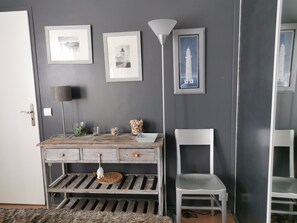 A room with a rustic wooden desk, a white chair, and a lamp. There are framed pictures on the wall and a small shelf with decorative items.