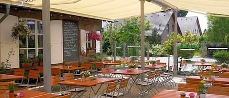 An outdoor café with wooden tables and chairs, a covered seating area, and a building with a chalkboard menu in the background.