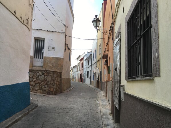 A narrow street with buildings on either side, featuring stone walls, metal bars on windows, and a street lamp.