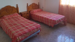 Ein Zimmer mit zwei hölzernen Betten, beide mit rosa karierten Bettdecken. Dazwischen steht ein kleiner Nachttisch aus Holz und auf der rechten Seite ein Fenster mit Vorhang.
