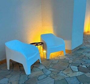 Deux chaises en plastique blanc, séparées par une petite table, sont placées sur un patio dallé de pierres, avec un mur en arrière-plan.