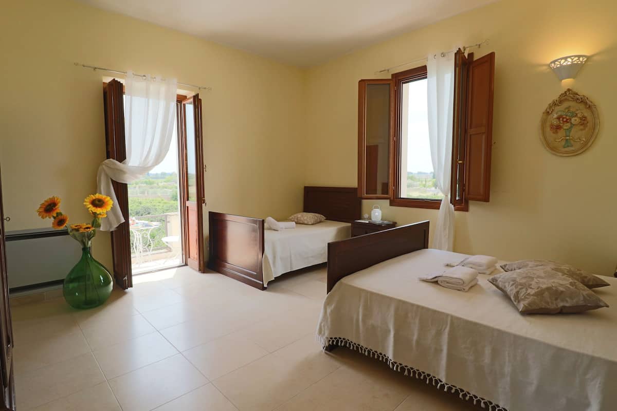Una habitación de hotel con dos camas, un jarrón verde con girasoles y un balcón con cortinas.