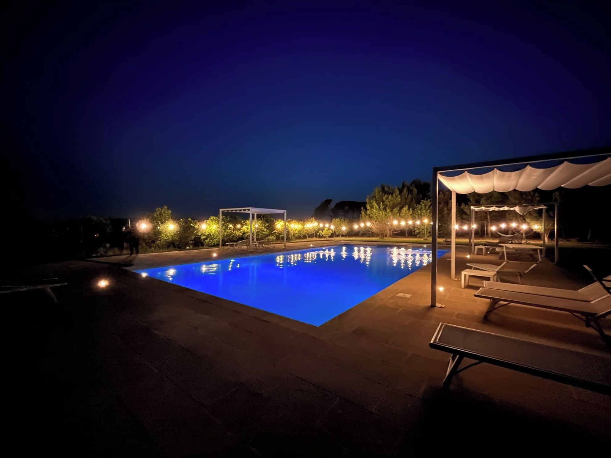 Una piscina illuminata di notte con superficie blu trasparente, circondata da sdraio e un'area salotto coperta.