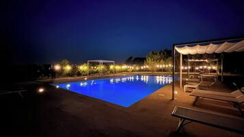 Una piscina illuminata di notte con superficie blu trasparente, circondata da sdraio e un'area salotto coperta.