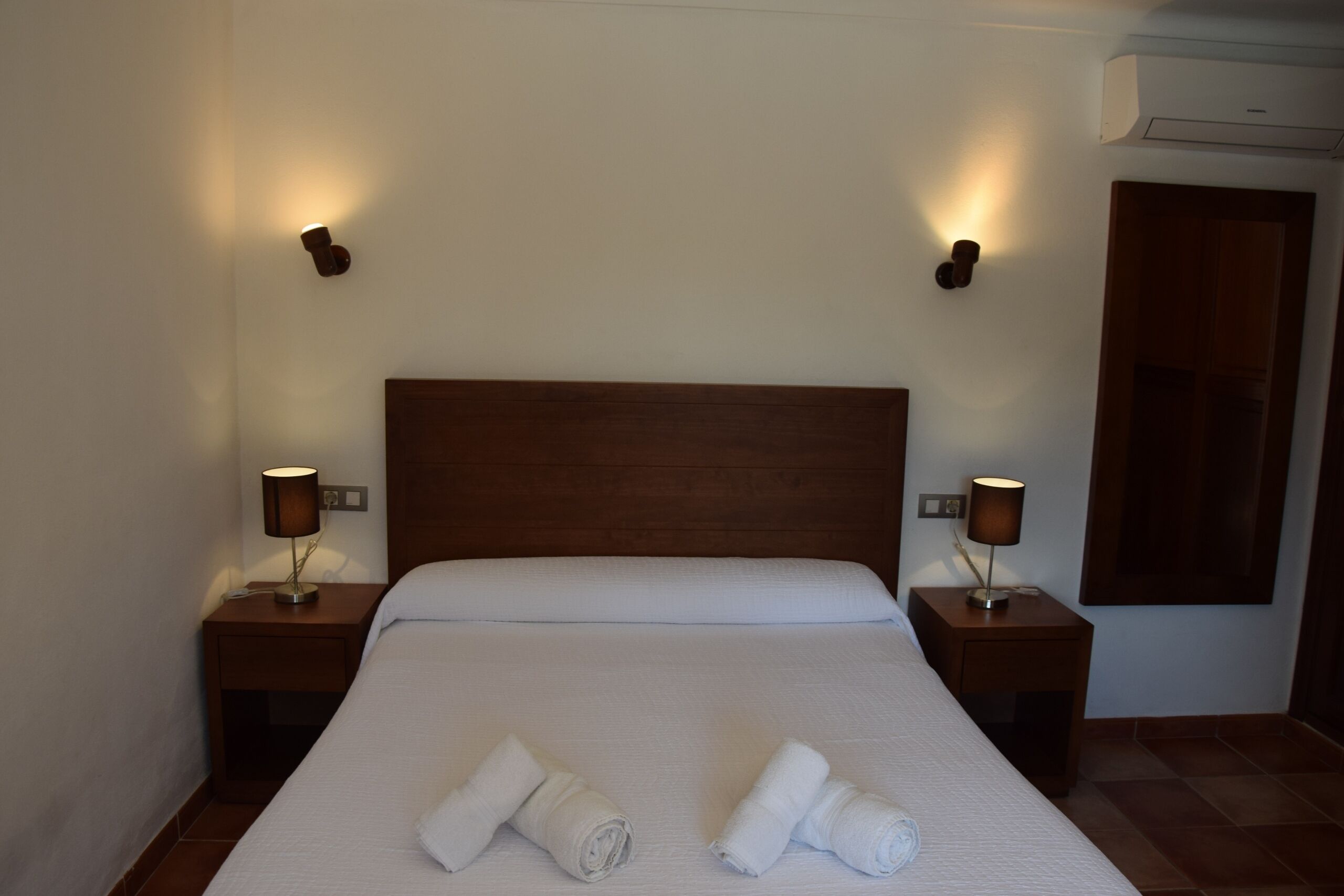 A neatly made bed with two rolled towels, two bedside lamps, and an air conditioning unit on the wall.