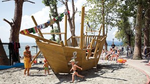 Una estructura de juego con forma de barco de madera en una playa arenosa, con árboles y vista al mar.