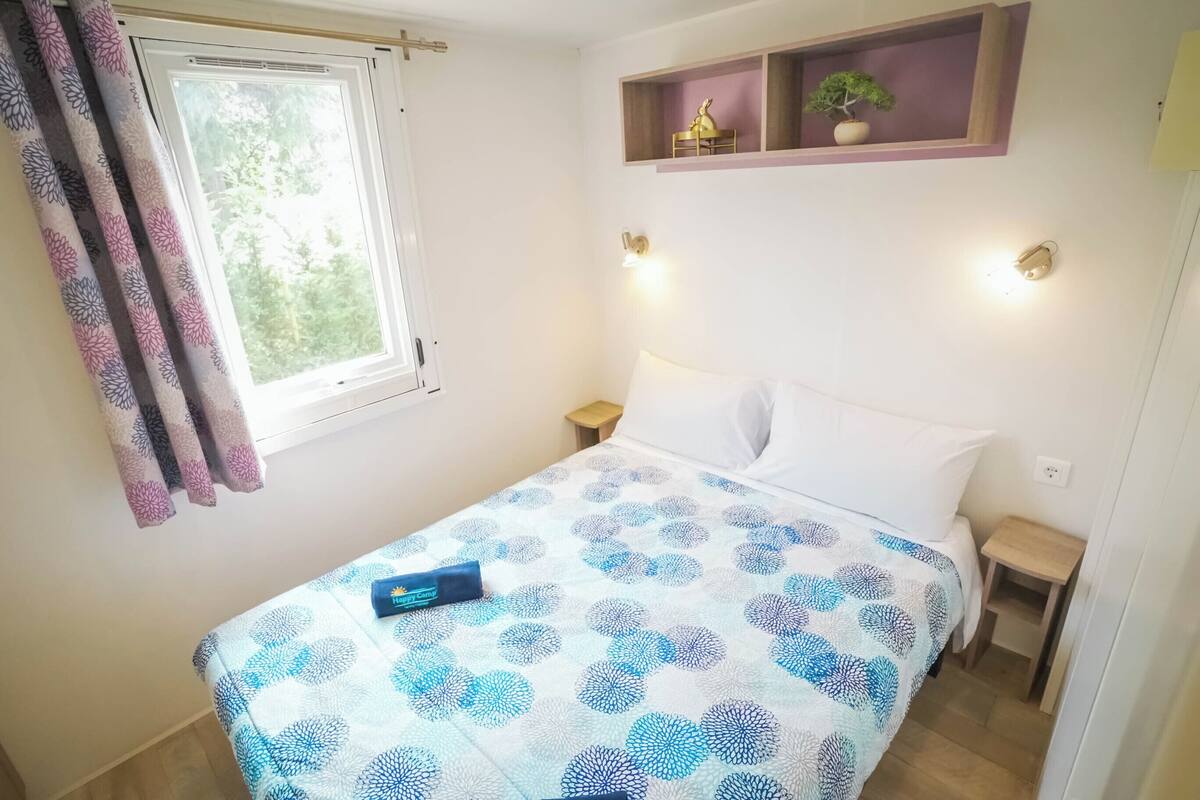 A neatly made bed with a patterned bedspread, a bedside table, a nightstand, a window with curtains, and a shelf with decorative items.