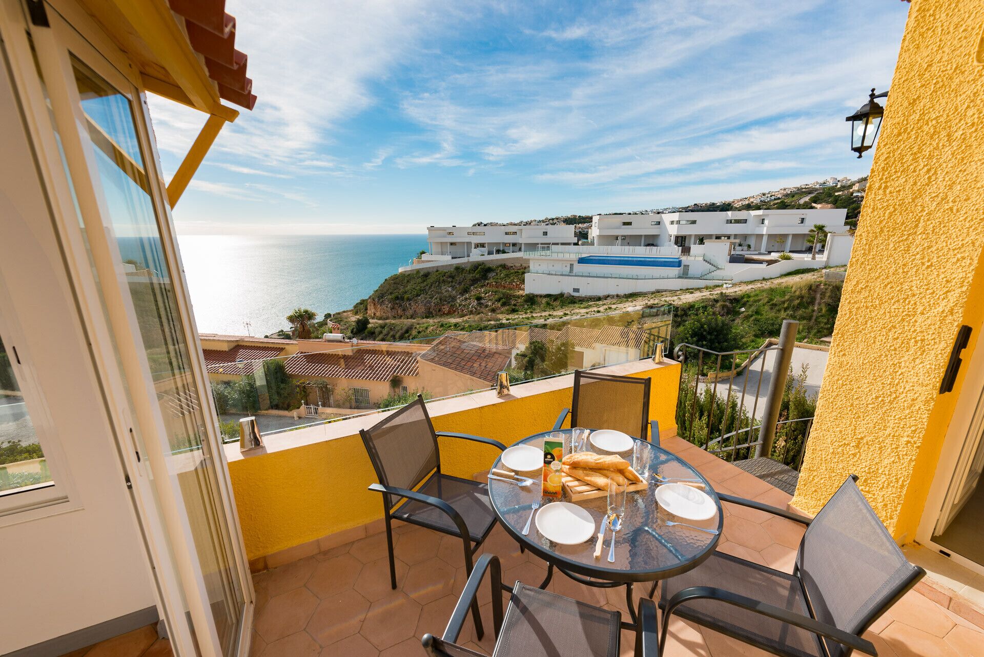 A balcony with a table set for two, overlooking a coastal view with buildings and a clear sky.