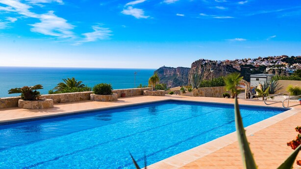 Una piscina rectangular con agua azul cristalina, rodeada de muros de piedra y plantas en macetas. Se divisa un acantilado rocoso con edificios en el fondo.