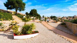 Un jardín ajardinado con un sendero, una maceta blanca con plantas y edificios de estilo mediterráneo al fondo.