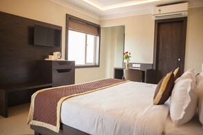 A hotel room with a large bed, a flat-screen TV mounted on the wall, a desk with a chair, a window with blinds, and an air conditioning unit.