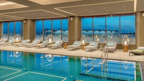 A modern indoor pool area with lounge chairs and a view of a cityscape at dusk.