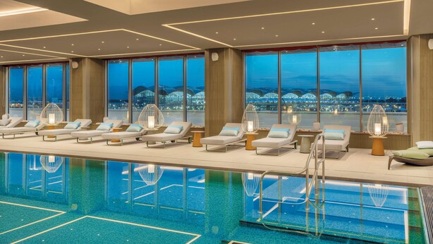 Un espace piscine intérieur moderne, doté de chaises longues et offrant une vue sur le paysage urbain au crépuscule.