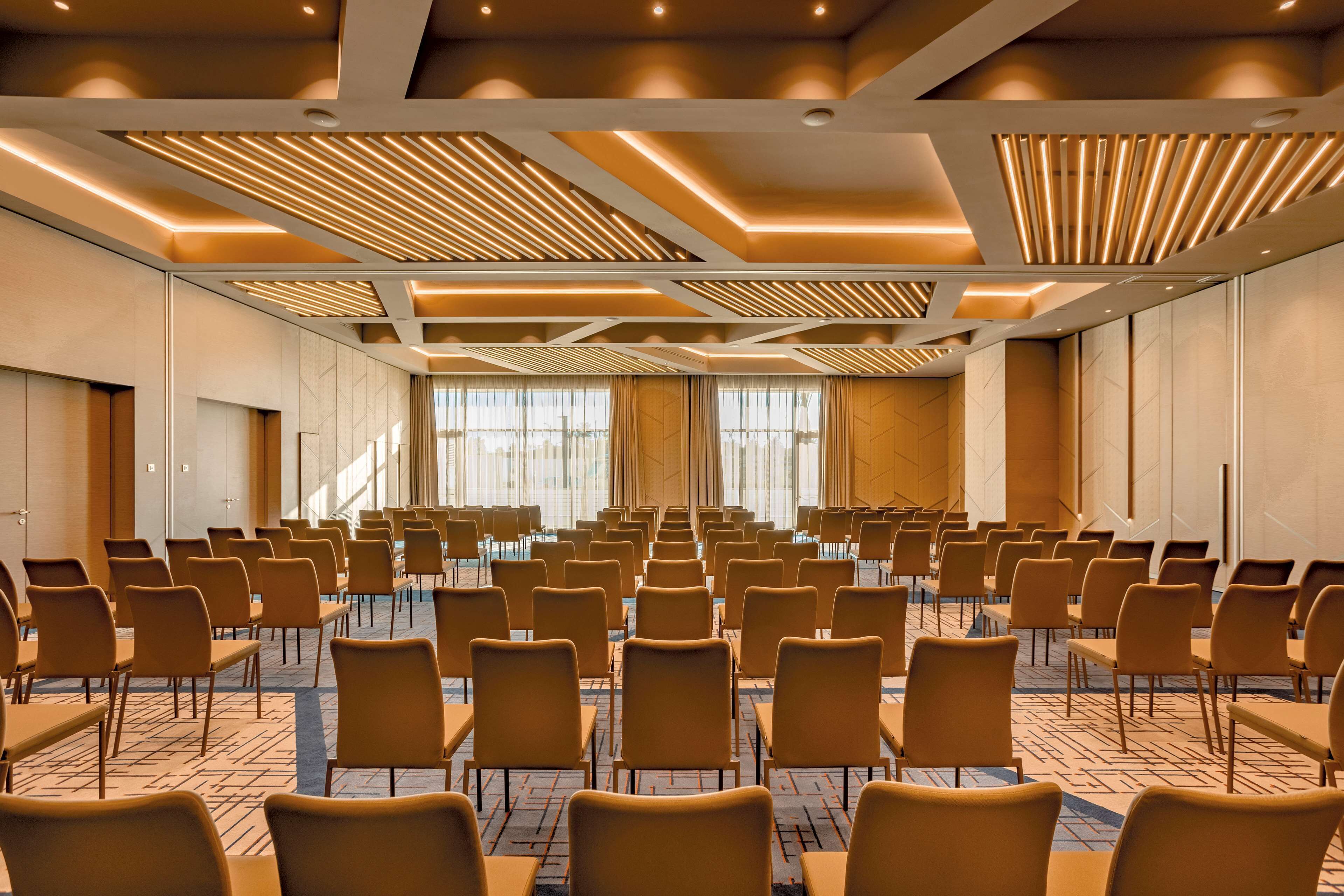Ein Konferenzraum mit Reihen von Stühlen und Tischen, großen Fenstern und modernem Deckenstil.