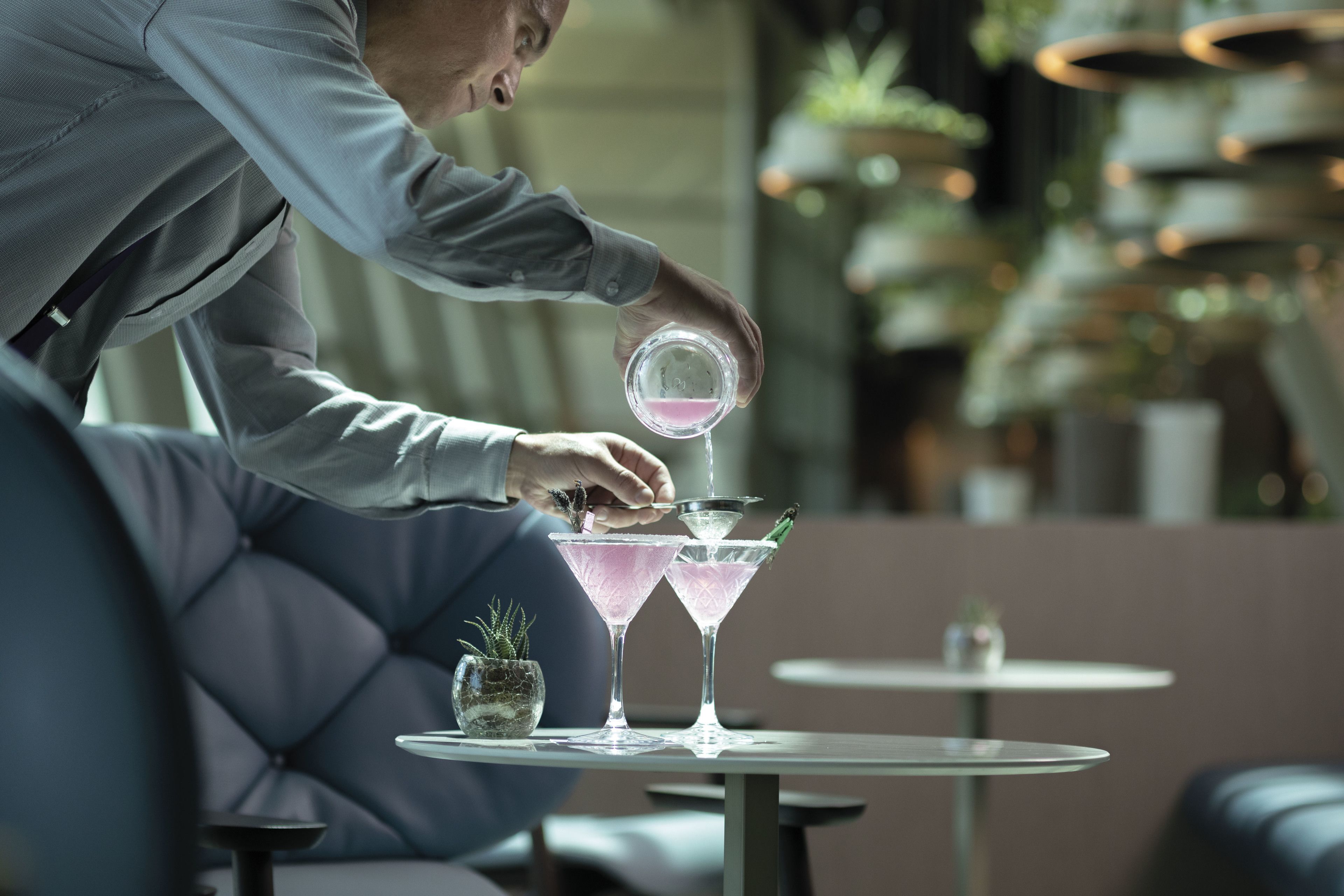 Una persona sirve una bebida en una copa de martini sobre una mesa, en un interior moderno con un sofá capitoné e iluminación colgante.